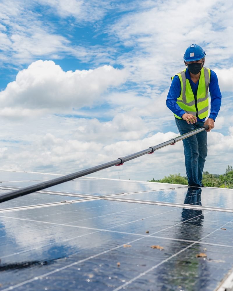 Professional worker cleaning solar panels with brush and washing with water on roof structure of building factory. Technician using mop to clean the dirty and dust, green electricity energy technology