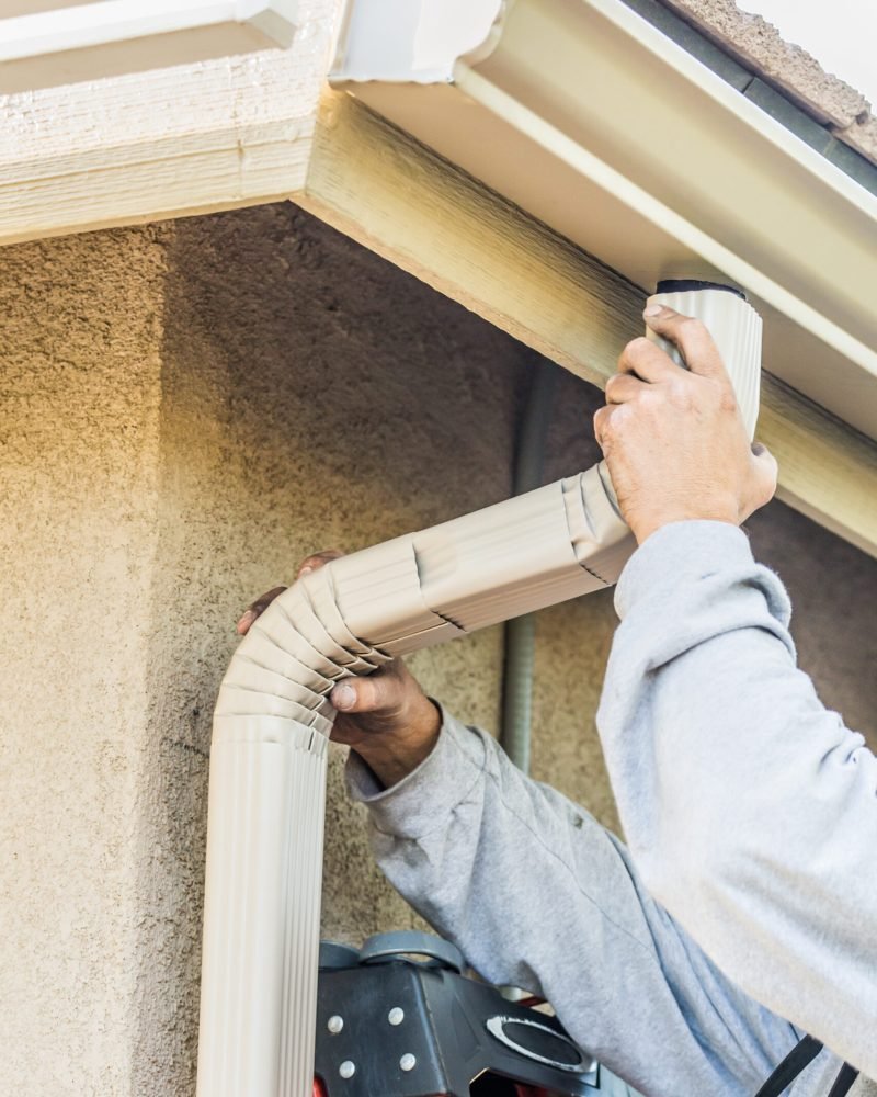 Worker Attaching Aluminum Rain Gutter and Down Spout to Fascia of House.