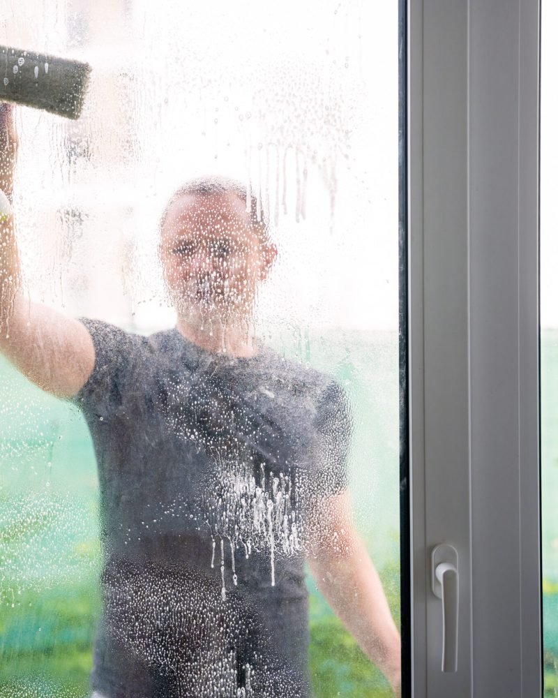 Young man cleans the windows in the house. He splashes detergent, rubs and removes water at the glass. High quality photo